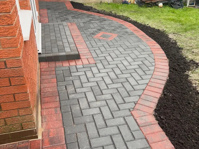 Charcoal herringbone driveway between a side gate and timber sleeper bed, surrounded by a two-course terracotta-red block border up to a white door and orange-tiled porch.