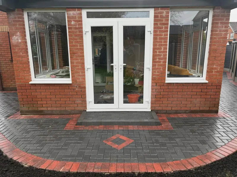 Charcoal herringbone patio and garden path with red brindle soldier border, channel drain across the width, recessed manhole and lawn alongside a brick house.