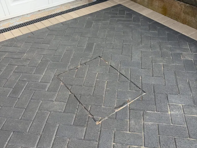 Rear charcoal herringbone patio with two flush manhole covers, red border and channel drain along the house, continuing as a path beside a green lawn and slatted fence.