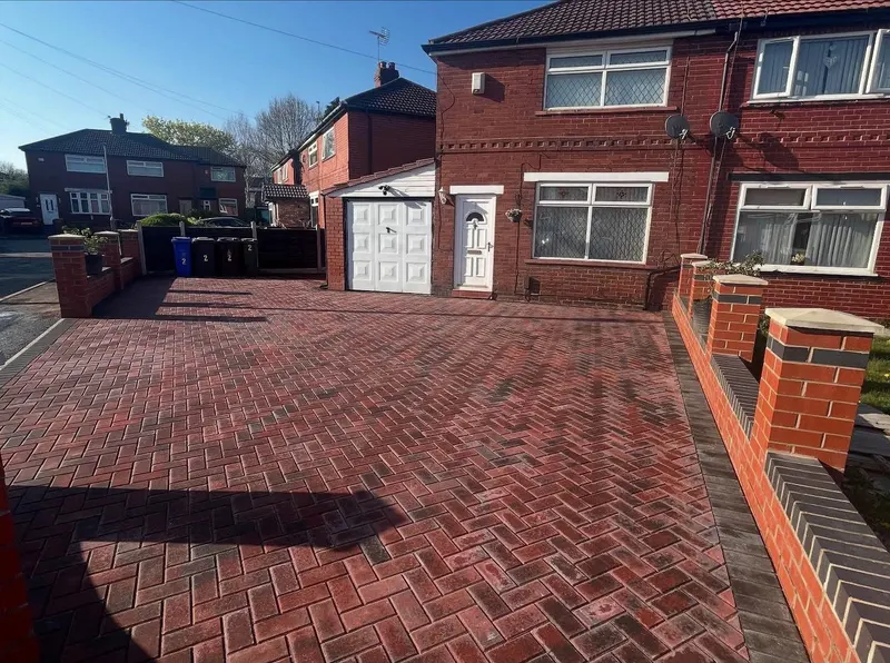 Detail of charcoal herringbone paving with cream double border, charcoal transition row, channel drain at a white garage threshold, and manhole infilled in herringbone.