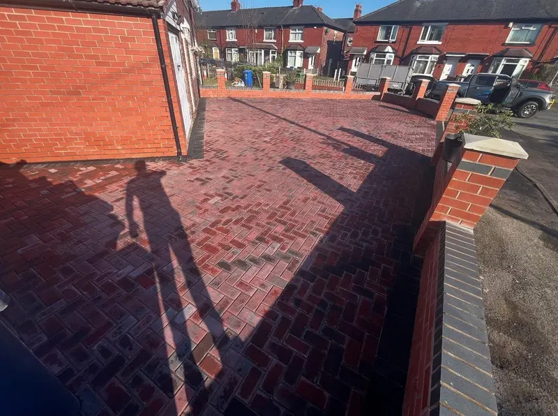 Warm red herringbone block driveway before white door and garage on a red brick semi, with charcoal border matching dark coping on new brick perimeter walls.