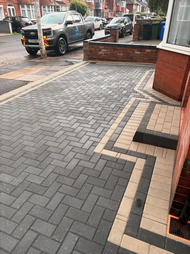 Charcoal herringbone paving before a white door with alternating charcoal and buff border courses, buff-topped step with charcoal riser, and flush manhole in the drive.
