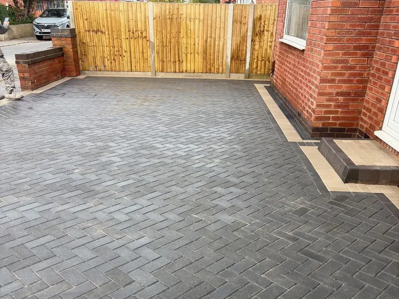 Charcoal herringbone driveway and entrance path with tan and charcoal layered borders, matching tan-tread step, and brick wall with dark coping along the side.