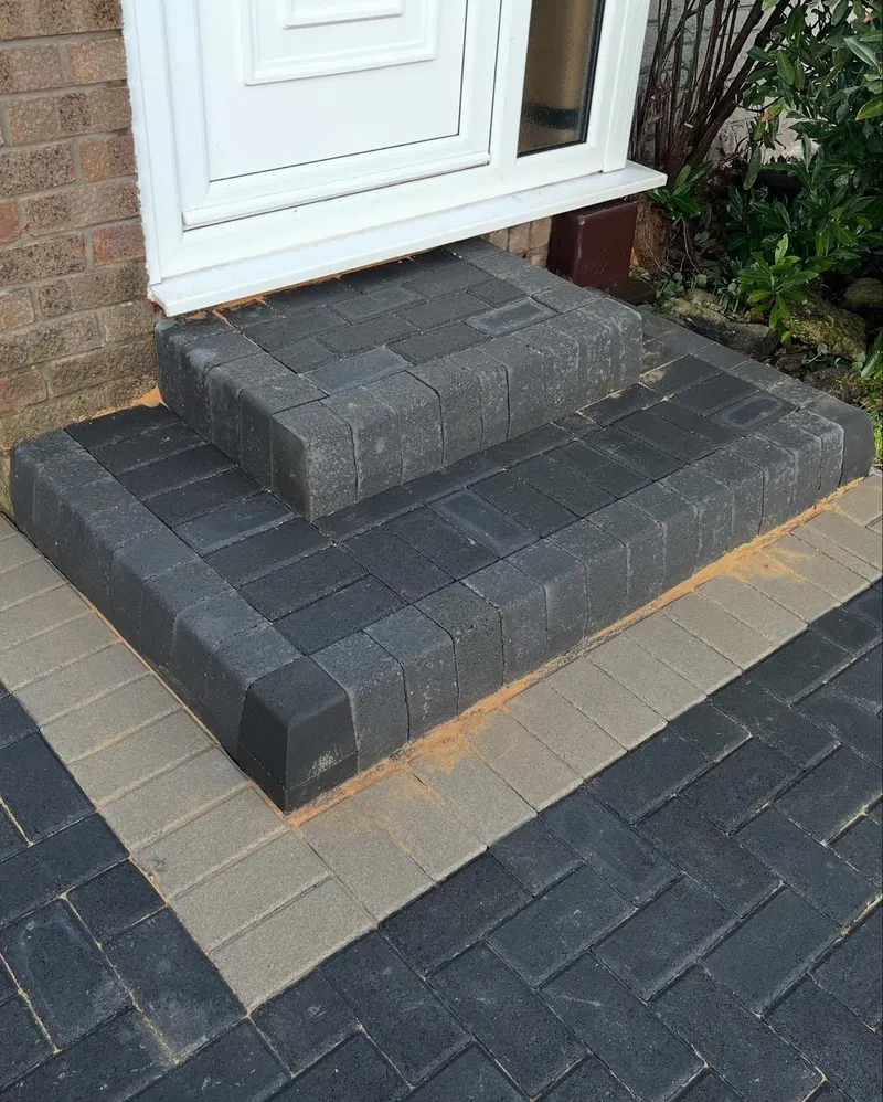 Charcoal herringbone block paving framed by a buff border, leading from the road past a wooden gate toward a white front door.
