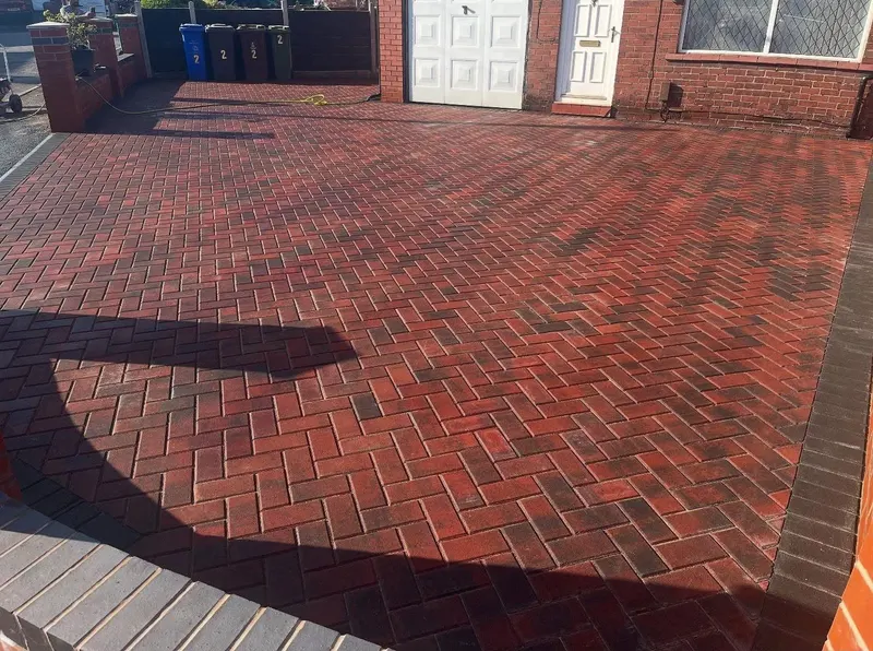Narrow side driveway of dark block paving in herringbone with a light border and linear drain, between brickwork and a wooden fence.