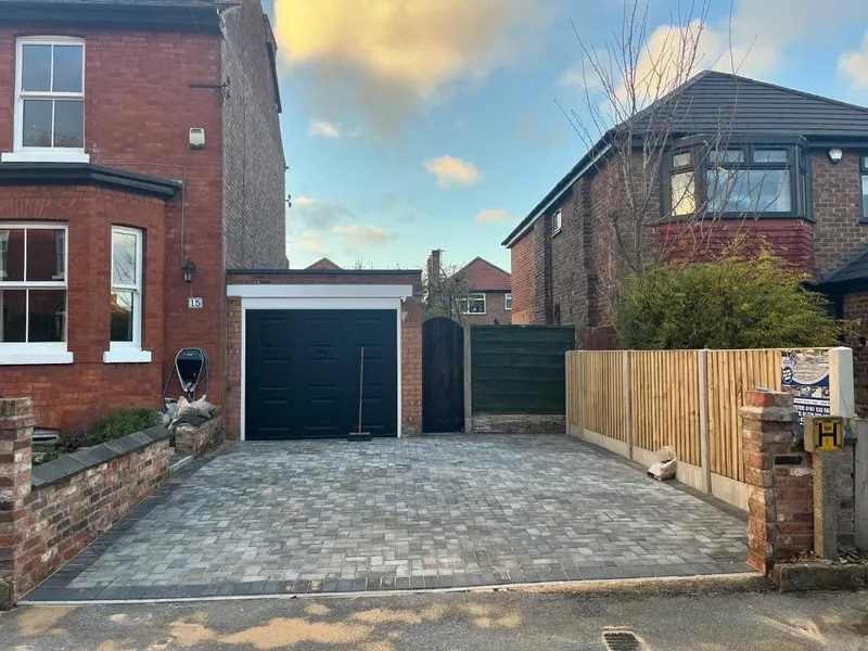 Slate-grey herringbone block paving with alternating light and dark border courses, curving around planting in front of a brick home.