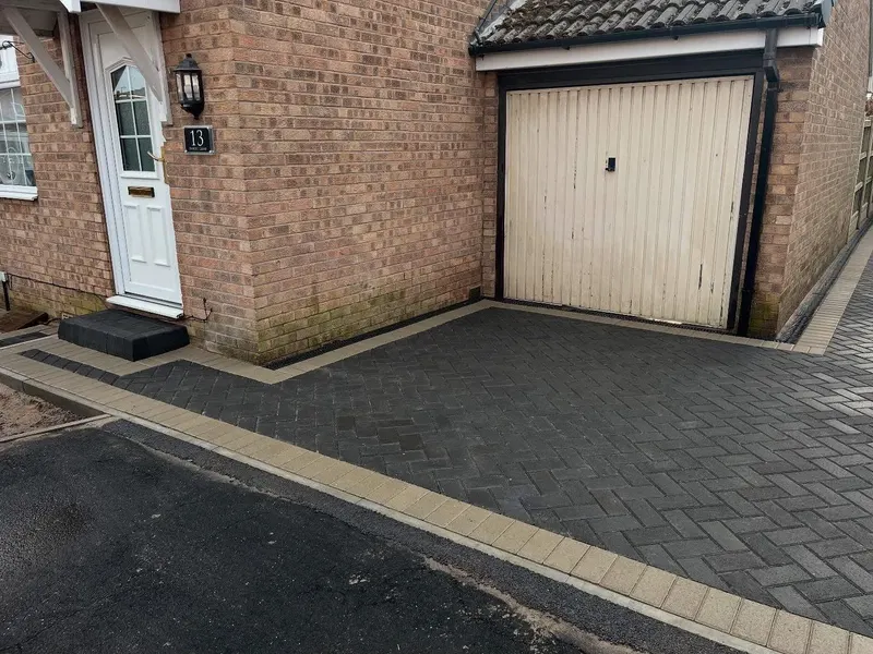 Multi-shade grey block paving in herringbone with a charcoal perimeter, laid before a black garage door and new timber fence.
