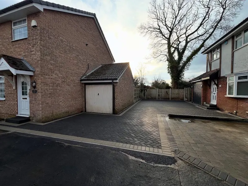 Dark herringbone block paving with a tan border linking a ribbed garage door and white front door on buff brickwork.