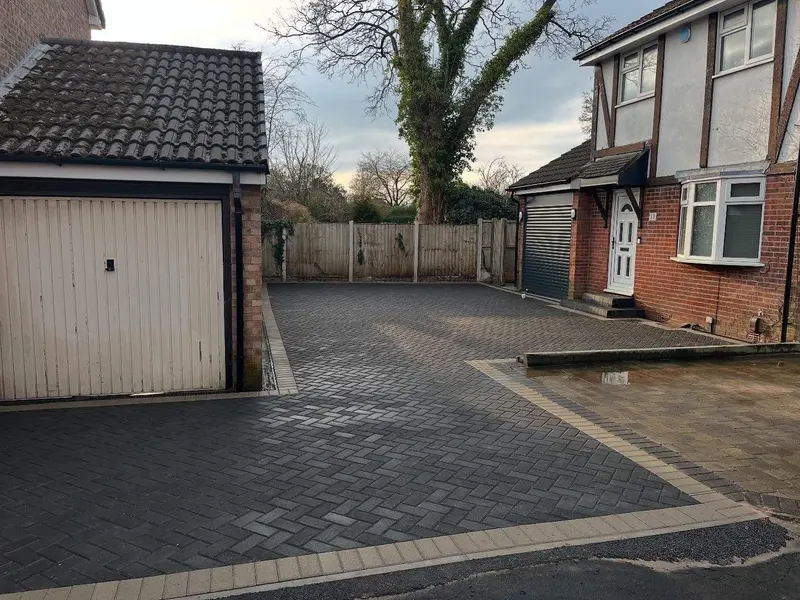 Rear garden paved in light grey herringbone block with a dark border, wrapping a central lawn and matching inset around a utility cover.