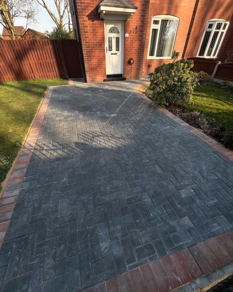 Charcoal herringbone block paving with tan double borders connecting a detached garage to the main house across a side courtyard.
