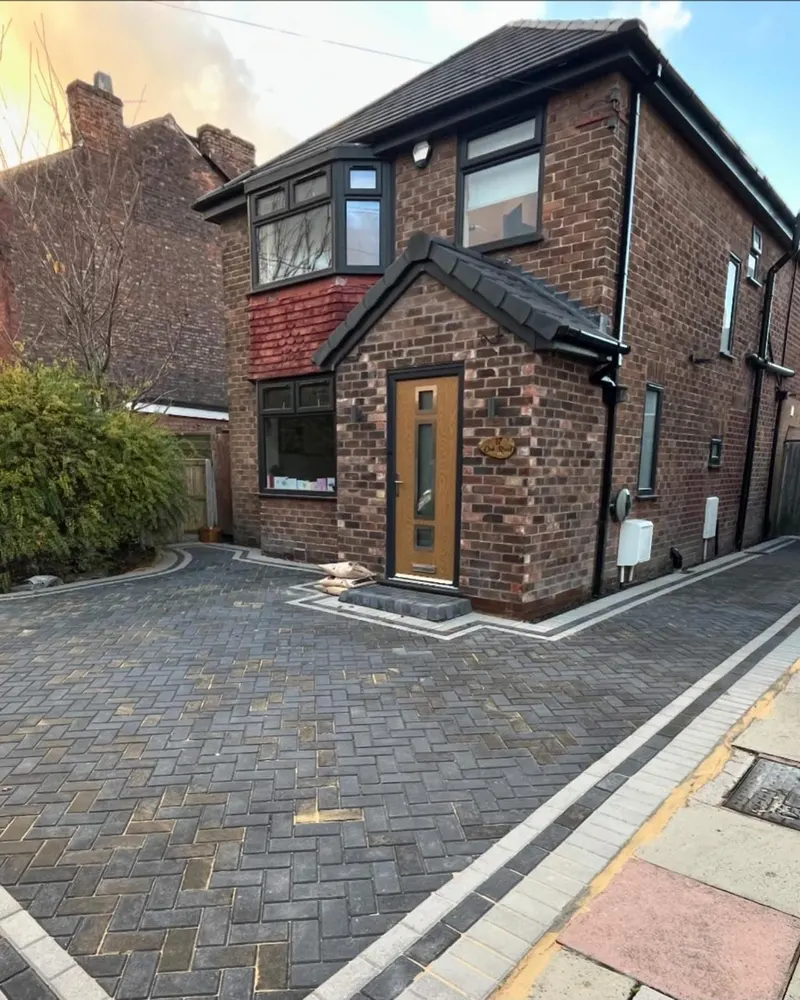 Front driveway of charcoal herringbone block paving framed by a reddish brindle soldier course, leading to a white door on a brick semi.