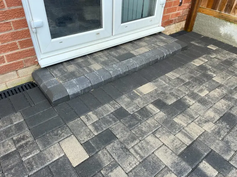 Charcoal herringbone block paving with layered grey and dark borders across the front and side path of a red brick house.