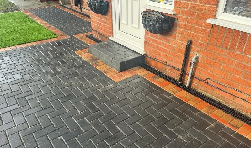 Smooth dark driveway centre edged in reddish block paving curves, flanked by green lawn panels in front of a brick detached house.