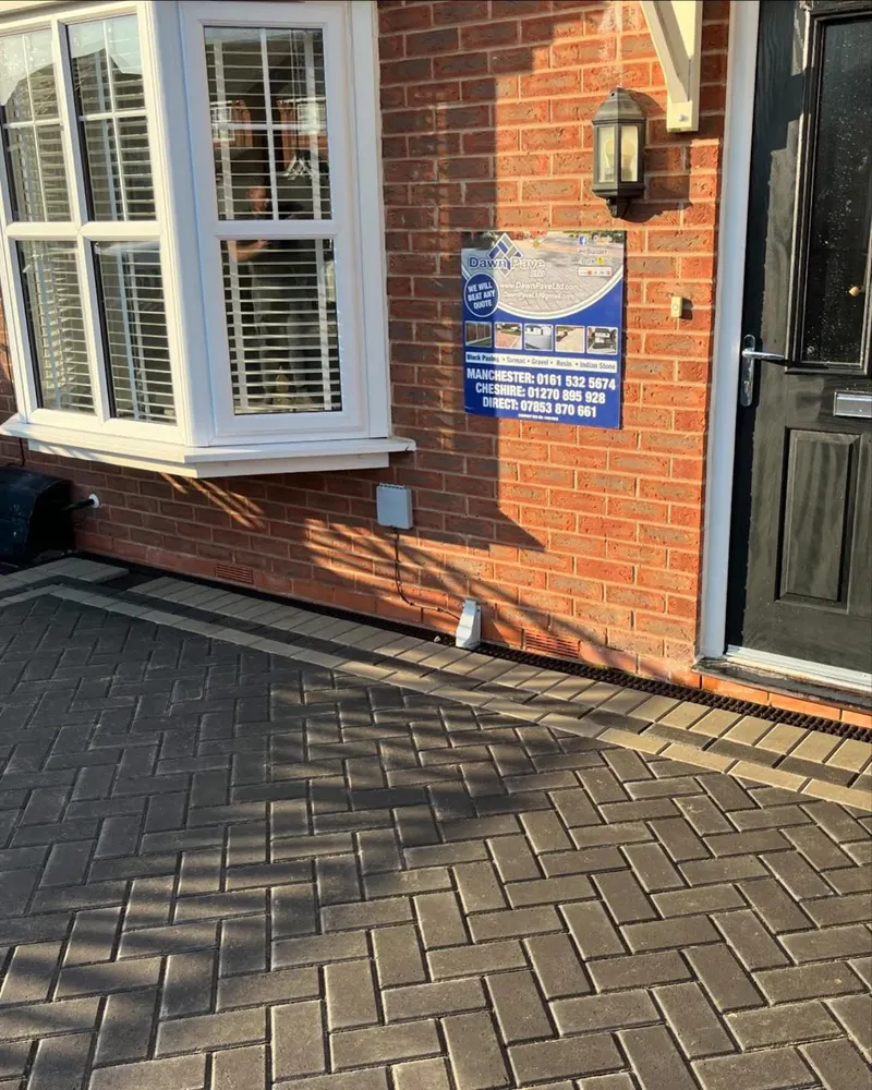 Charcoal herringbone block paving with grey stretcher borders, cut neatly around a metal pole and manhole between fence and brick wall.