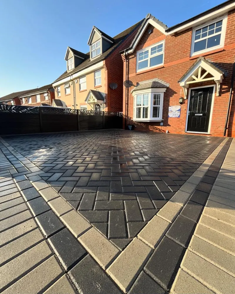Fresh charcoal herringbone block paving with sandy border courses along the base of a red brick house below a white bay window.