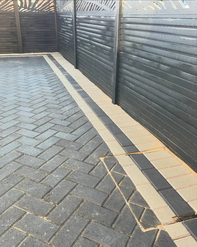 Low-angle view of charcoal herringbone block paving framed by alternating light and dark border bands in a front courtyard.