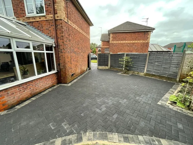 Grey-toned block patio with charcoal borders, a small step to a white door, slot drain and a paved-over recessed manhole.