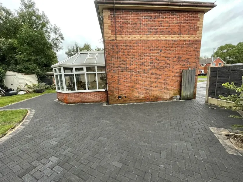 Charcoal herringbone block paving with a light grey border along a conservatory wall, fence and small planted tree.