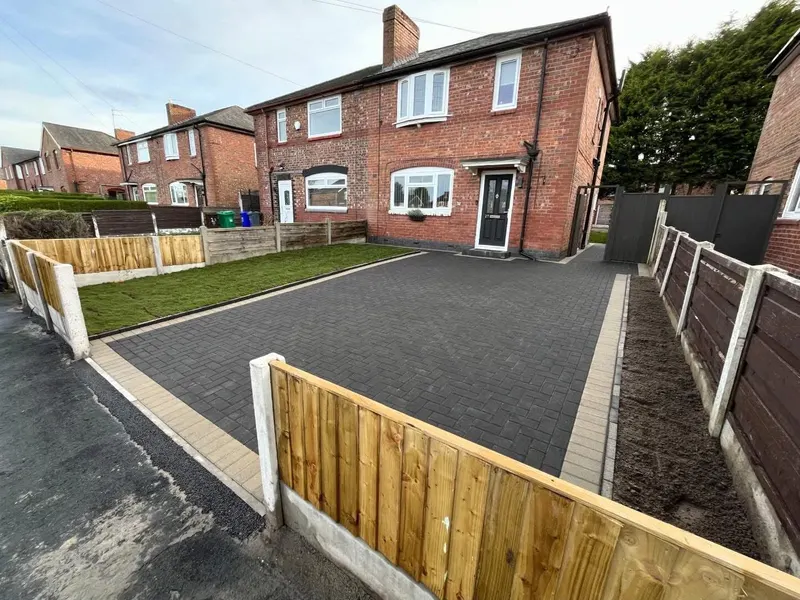 Charcoal herringbone block driveway edged in tan and dark header courses, opening through wrought iron gates from flagstone paving.