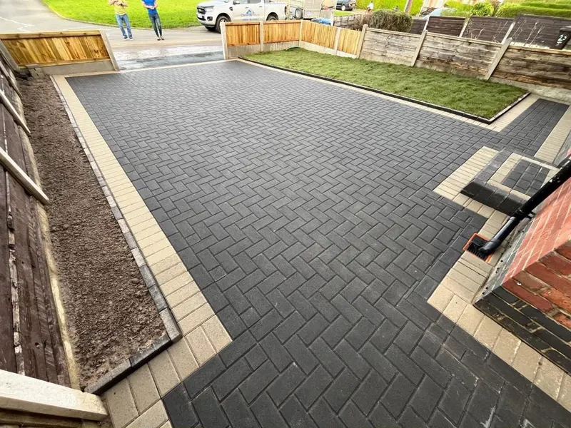 Dark grey staggered block paving with a buff border outlining a small front lawn, enclosed by new vertical-slat fencing.