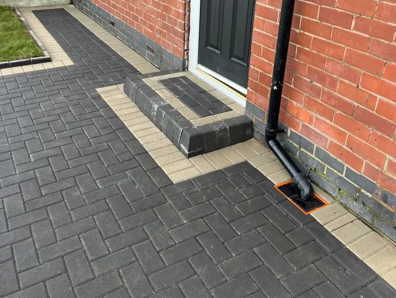 Charcoal herringbone block paving with a tan front edge, small lawn and fence, as contractor and homeowner shake hands.