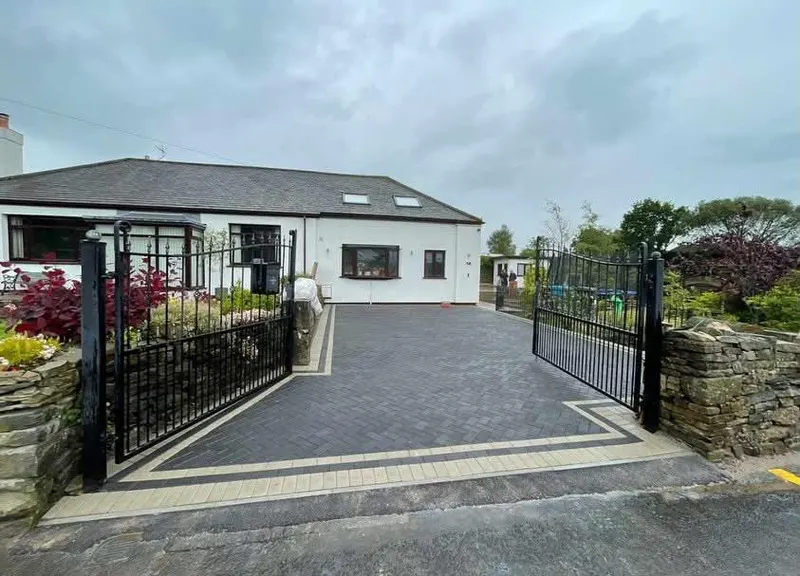 Charcoal herringbone block driveway bordered in light grey blocks, leading past lawn and hedge to a green front door.
