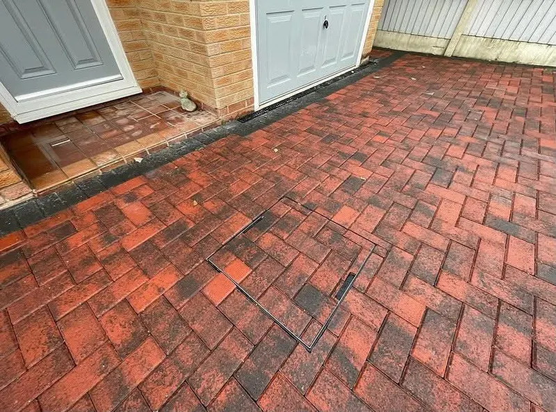 Wide charcoal herringbone block paving with decorative cream and dark borders, viewed through open ornamental gates toward a white bungalow.