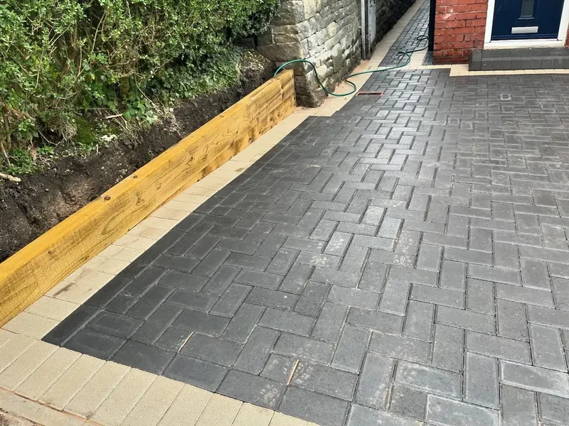 Full-width charcoal herringbone block driveway framed by a terracotta double border in front of a part-rendered brick house.