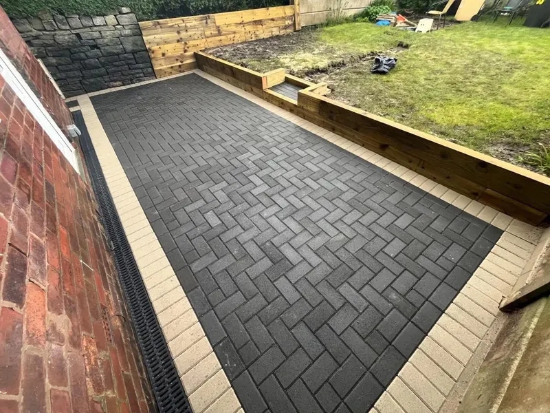 Charcoal herringbone block paving with tan double borders beside a timber sleeper retaining wall and stone boundary.