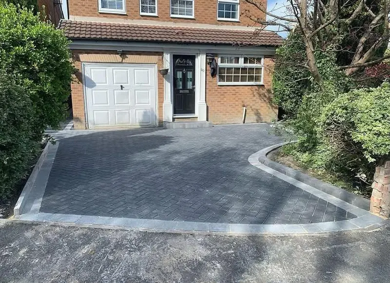Rectangular charcoal herringbone block patio with a cream border and channel drain, set against brick and sleeper walls.