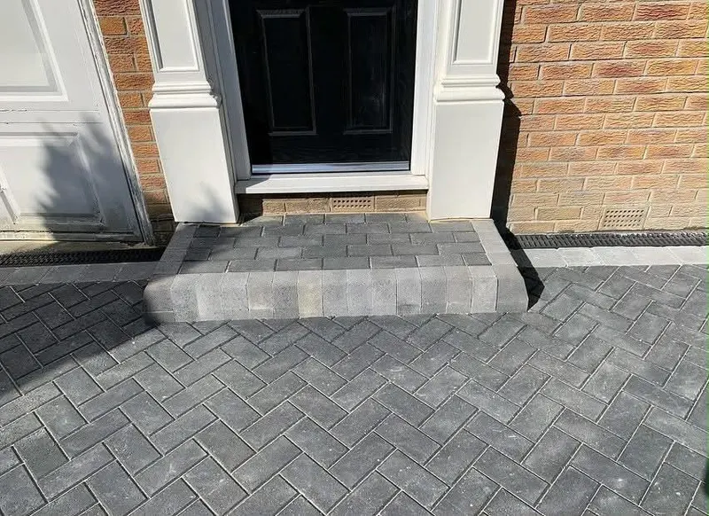 Dark herringbone block driveway with lighter grey edging that curves around a planting bed before a brick house and garage.