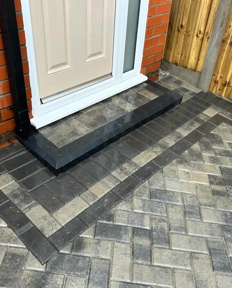 Multi-tonal grey herringbone block driveway glistening on a wet day, bordered along a wooden fence to the street.