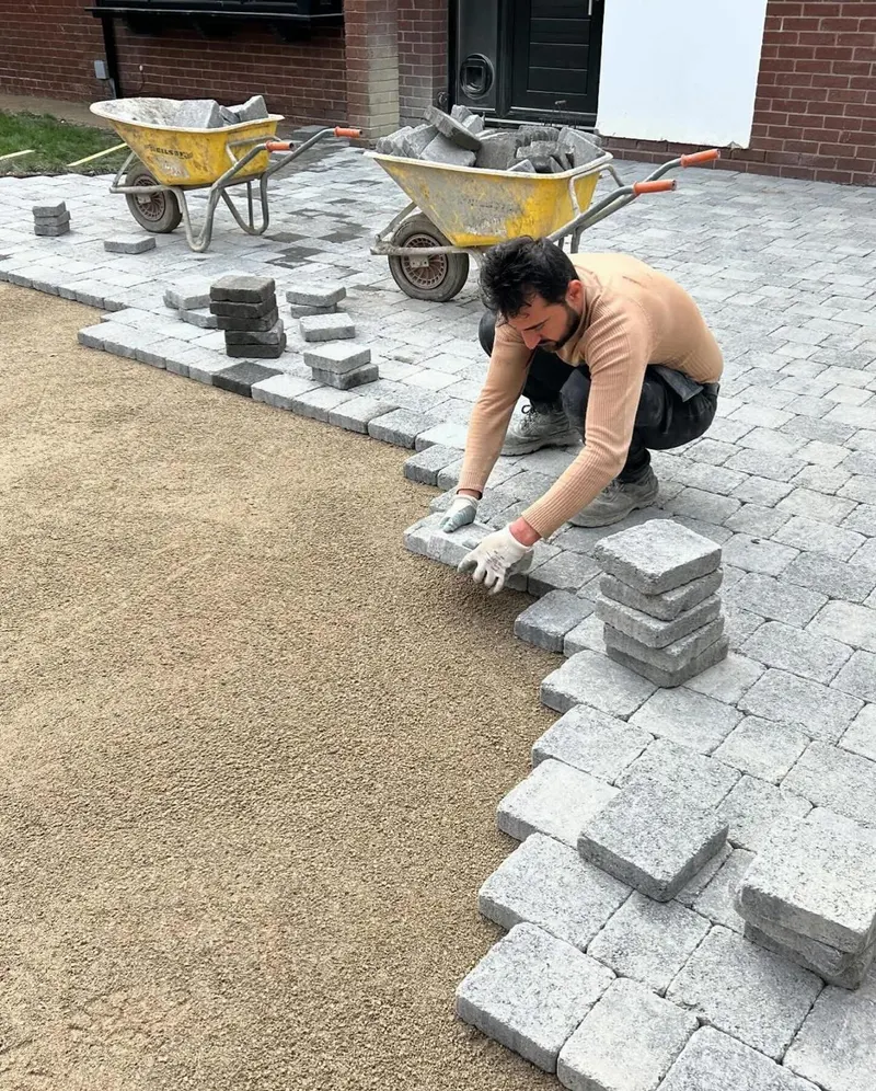 Pale grey tumbled block paving in running bond filling the front garden of a red brick house with black doors and garage.