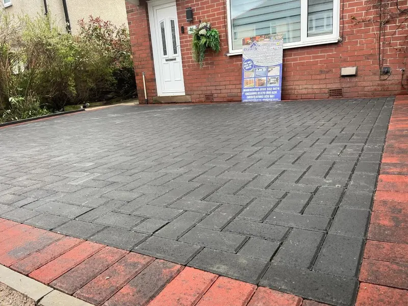Paving installer placing light grey tumbled block pavers on sand beside stacked blocks and wheelbarrows at a brick house.