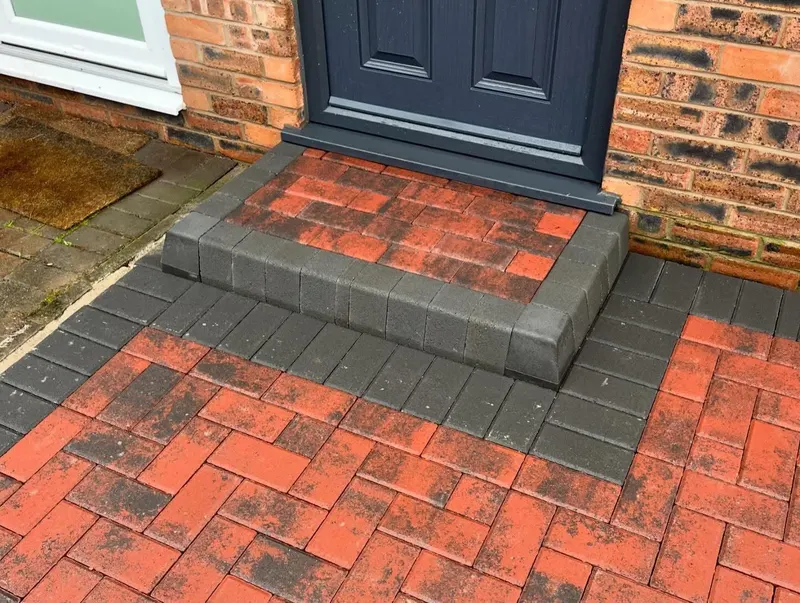 Charcoal herringbone block driveway surrounded by a terracotta double border, set before red brick with shrubs to one side.