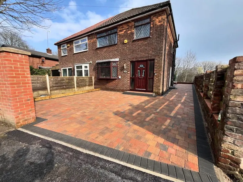 Narrow brindle herringbone block walkway with charcoal soldier edges and a recessed manhole paved to match, between brick and fence.