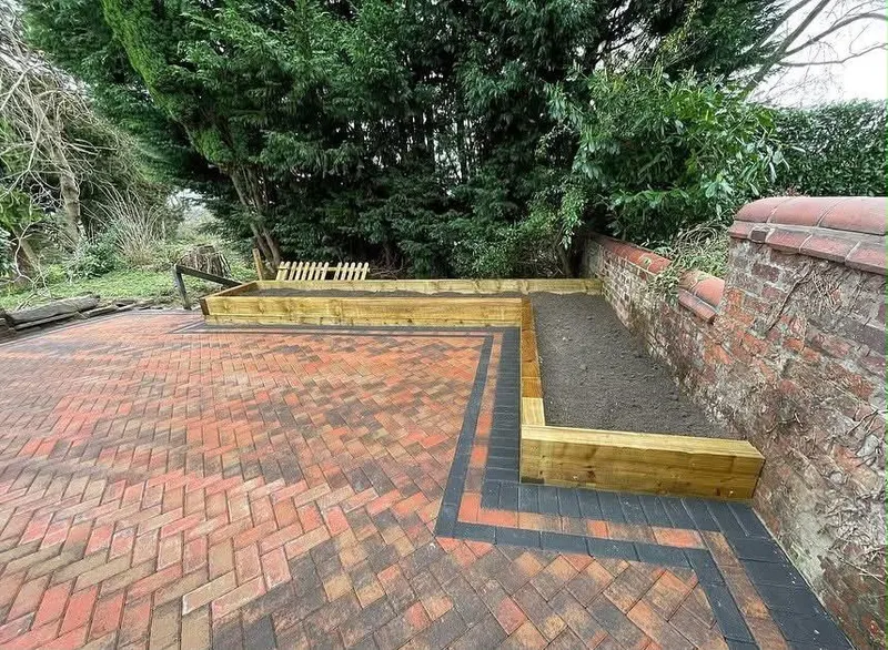 Red-toned herringbone block paving edged in charcoal, opening toward timber sleeper raised beds and a large brick house.