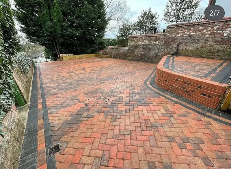 Red herringbone block paving with charcoal borders surrounding L-shaped timber sleeper planters beside an old brick wall.