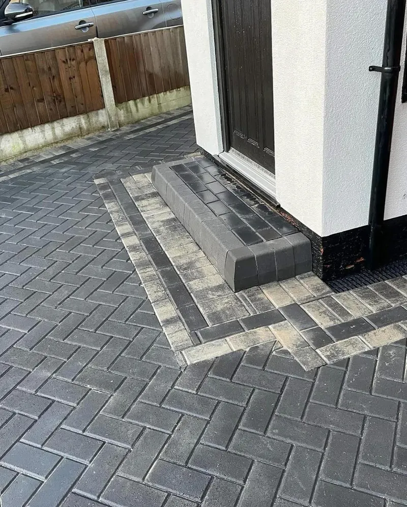 Expansive red and grey herringbone block paving with dark borders, inlaid manhole and raised brick planter in a walled garden.