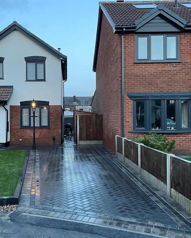 Charcoal herringbone block paving with cream and dark triple borders framing a paved step at a white-rendered wall.