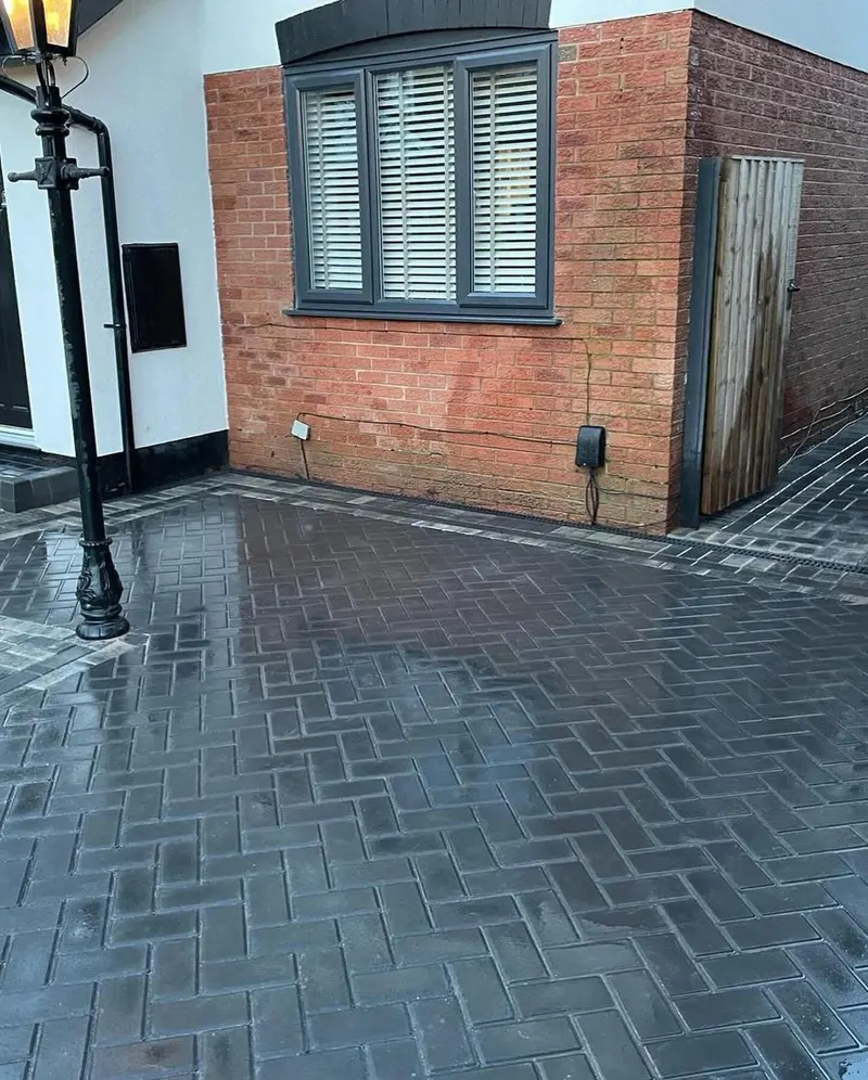 Glossy charcoal herringbone block paving with grey borders running between two houses, slot drain and lamp post visible.