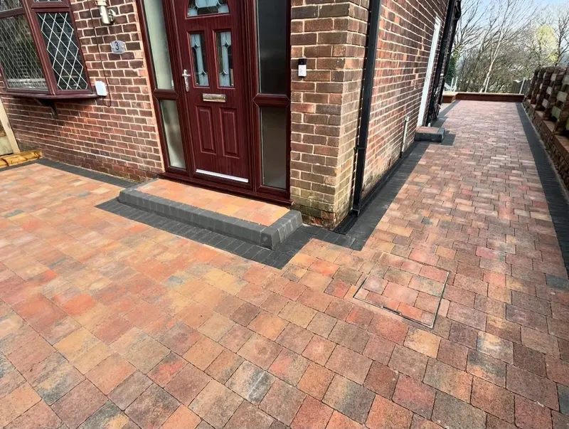 Autumn-mix brindle block paving with charcoal borders, recessed manhole tray and small matching step beside a brick wall.