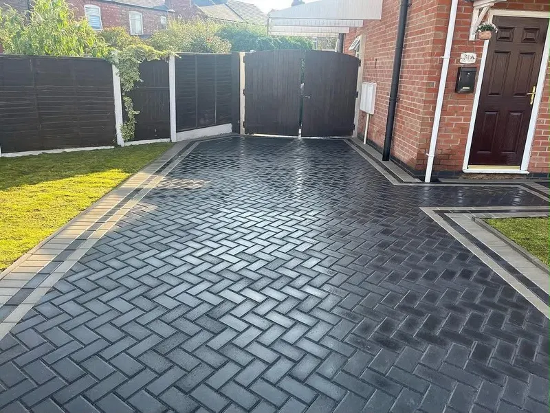 Red and grey brindle herringbone block paving with a charcoal double border meeting the kerb in front of a brick house.