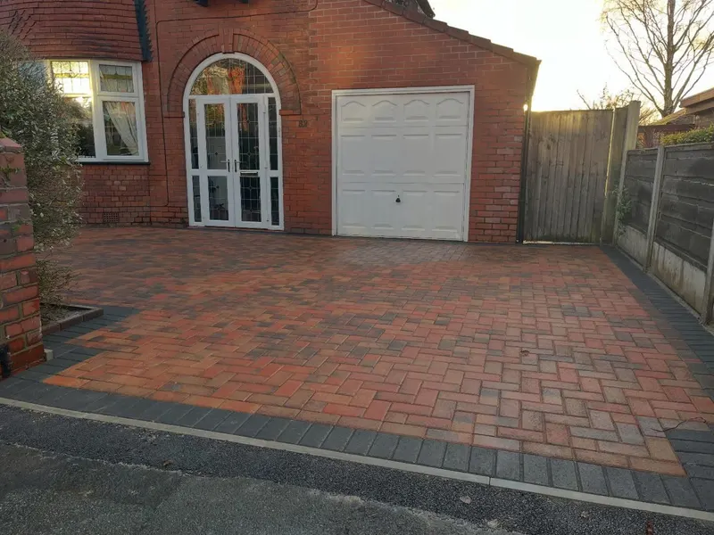 Charcoal herringbone block paving with grey and dark border bands and a recessed utility cover finished in matching blocks.