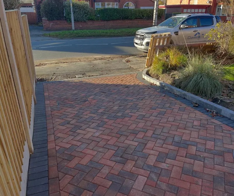 Red brindle herringbone block paving on a gentle slope, charcoal border, wooden fence and gate under a clear sky.