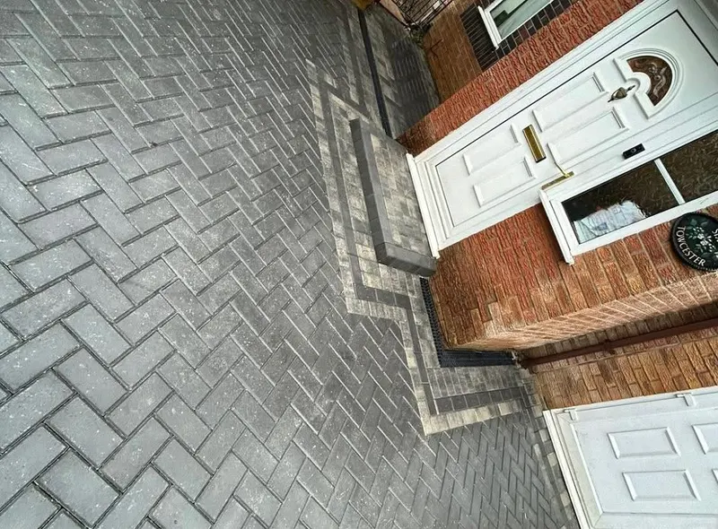 Charcoal herringbone block paving with buff borders and two manholes framed in matching blocks at a brick bungalow entrance.
