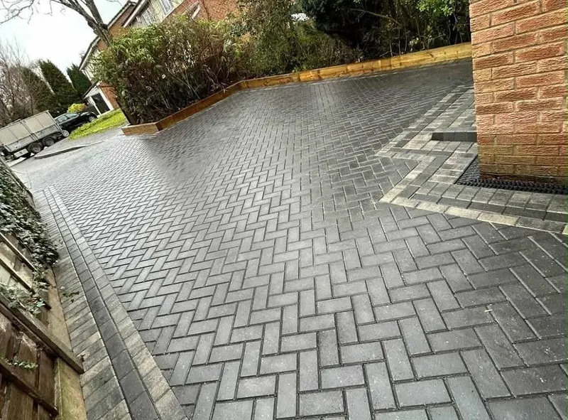 Charcoal herringbone block paving with alternating light and dark borders, bullnose step and slot drain at white doors on brick.