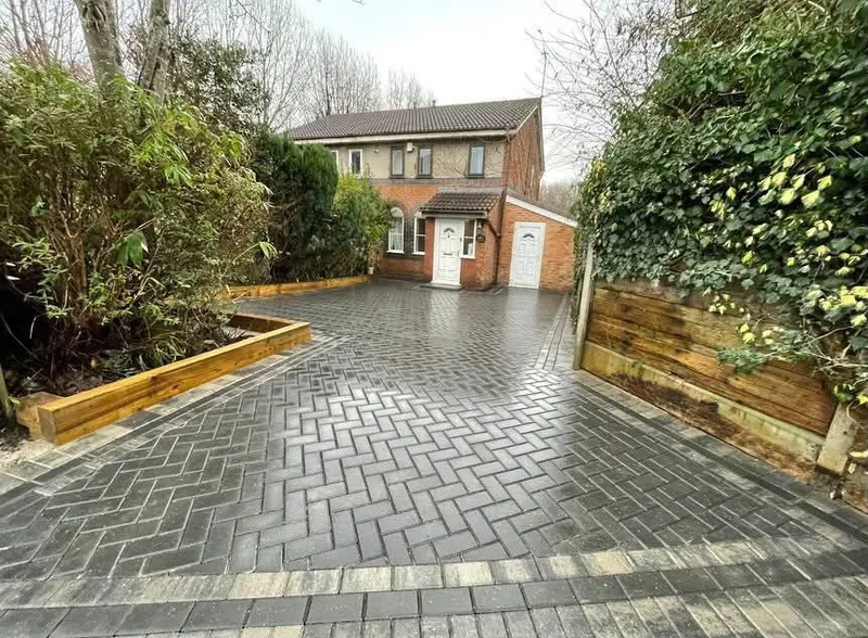 Spacious charcoal herringbone block driveway edged in silver-grey double borders, stepping up toward a brick wall and sleeper beds.