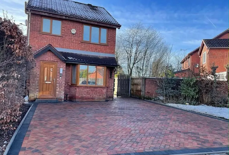 Charcoal herringbone block paving with light borders leading to a red brick house, timber sleeper planters and ivy-covered fence.