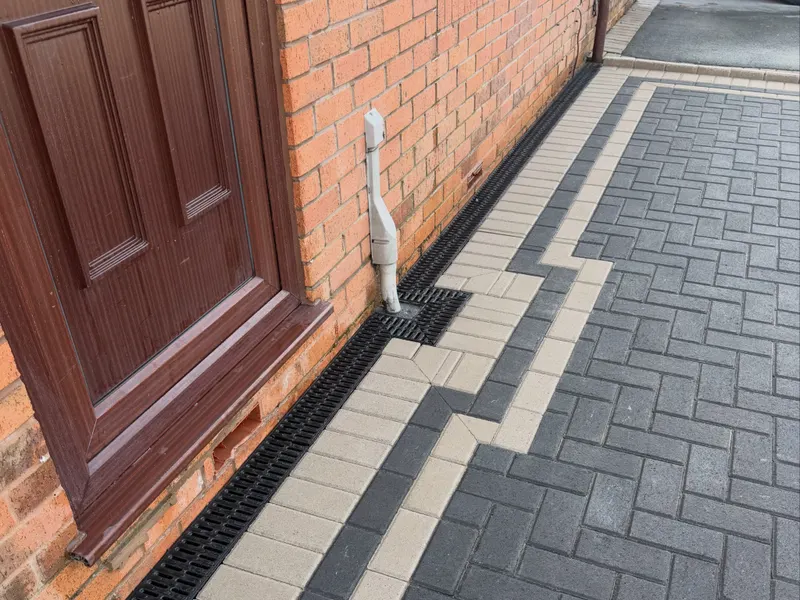 Reddish herringbone block paving with charcoal border running past new fence panels toward a rear patio with outdoor seating.